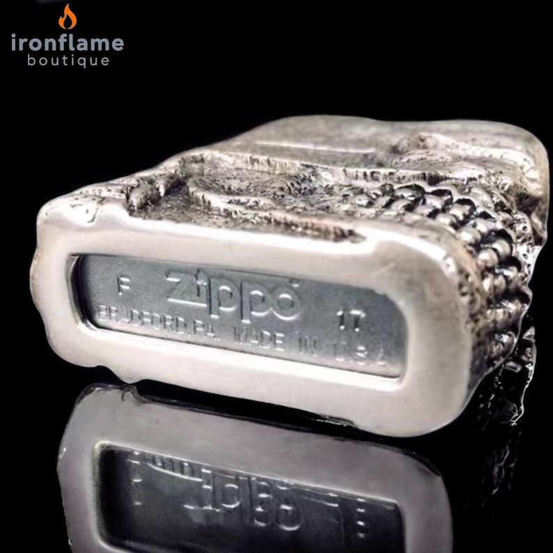 May include: A silver Zippo lighter with a textured, rugged design. The lighter's base features the Zippo logo and the text "BRADFORD, PA. MADE IN U.S.A." The lighter is reflected on a black surface.