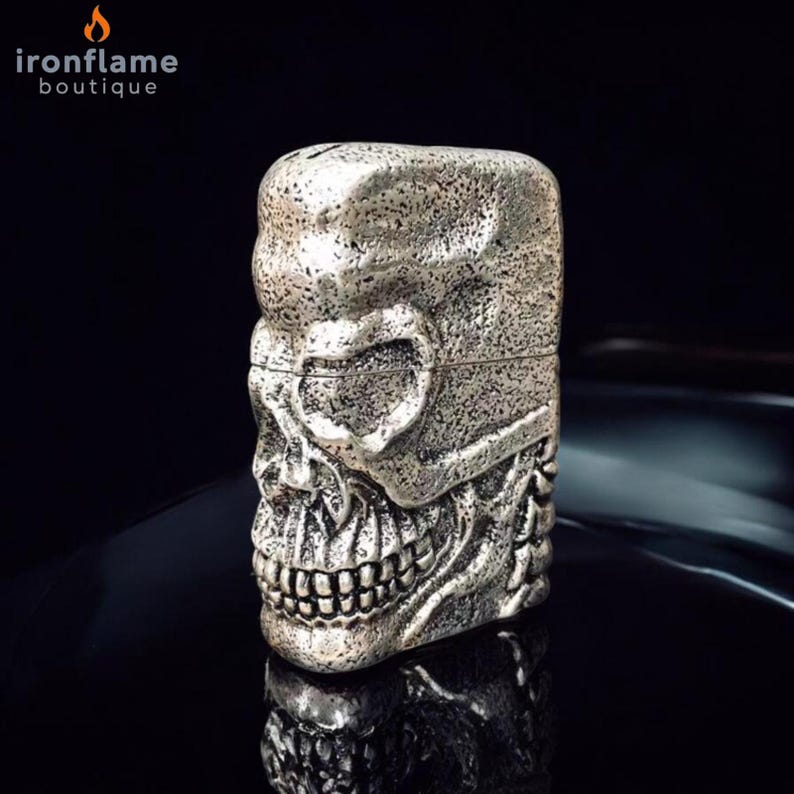 May include: A silver-coloured skull-shaped lighter with detailed facial features, including eye sockets, a nose cavity, and teeth. The lighter has a textured surface and the brand name "ironflame boutique" is visible in the upper left corner.