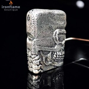 May include: A silver skull-shaped lighter with a textured finish. The lighter features a hinged lid and detailed skull features, including teeth and eye sockets. The brand name "ironflame boutique" is visible in the top left corner.