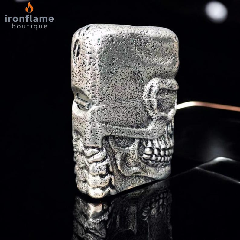 May include: A silver skull-shaped lighter with a textured finish. The lighter features a hinged lid and detailed skull features, including teeth and eye sockets. The brand name "ironflame boutique" is visible in the top left corner.