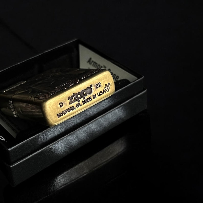 May include: A gold-colored Zippo lighter with intricate engraved designs, resting in a black box. The lighter's surface reflects light, and the text "Zippo 22" and "BRADFORD, PA. MADE IN USA" are visible.