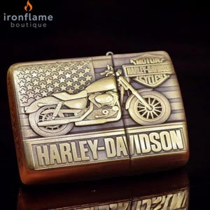 Pu&ograve; includere: Un accendino rettangolare color bronzo con un design di motocicletta Harley-Davidson. L'accendino presenta un motivo a bandiera americana con stelle e strisce, e il logo Harley-Davidson &egrave; ben visibile.