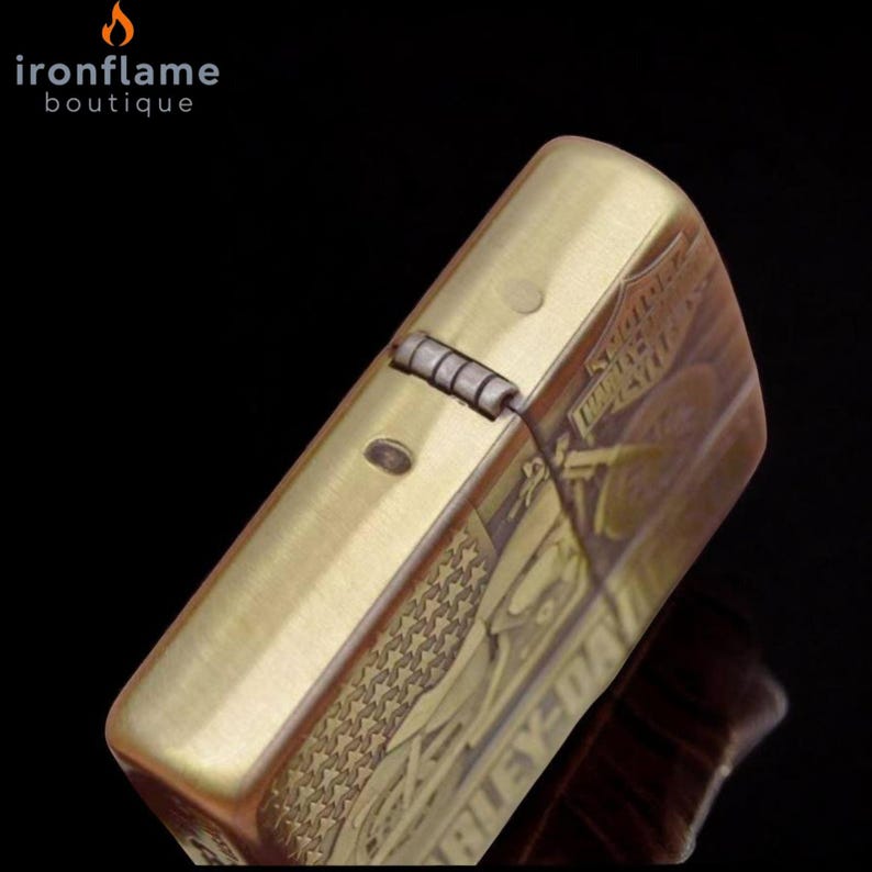 Pu&ograve; includere: Un accendino color ottone con coperchio incernierato. L'accendino presenta dettagli incisi, tra cui stelle e la scritta "HARLEY-DAVIDSON". Il logo "ironflame boutique" si trova nell'angolo in alto a sinistra.