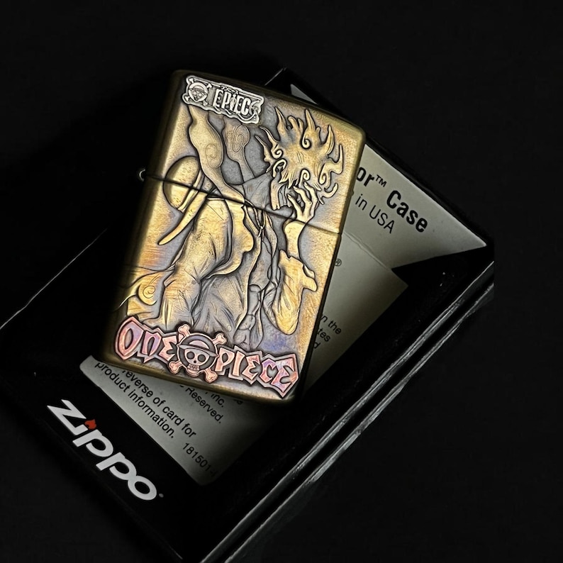 May include: A brass-colored Zippo lighter with a detailed embossed design featuring a character from the "One Piece" anime series. The lighter has the text "ONE PIECE" and "EPIEC" on it. The lighter is in its original black box.
