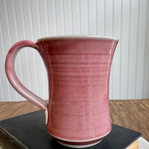 May include: A handmade, tall ceramic mug in a dusty rose color. The mug has a wide, flared shape with a rounded handle and a white rim. The mug is sitting on a dark book.