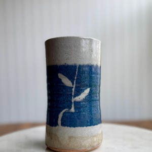 May include: A cylindrical ceramic vase with a blue band featuring a white leaf design. The top and bottom sections are a light gray color. The vase is handmade and has a rustic, artisanal aesthetic.