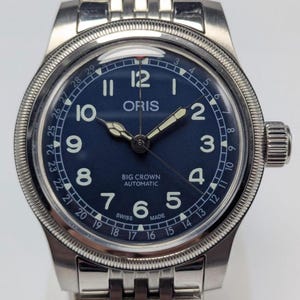 May include: A stainless steel Oris Big Crown Automatic watch with a dark blue dial. The watch features white numerals, hands, and a date display. The watch band is a stainless steel link bracelet. The watch is Swiss made.