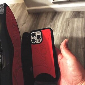 May include: A red and black phone case with a logo design. The case is held in a hand, with a black shoe with a red sole visible in the background.
