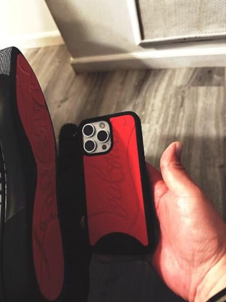 May include: A red and black phone case with a logo design. The case is held in a hand, with a black shoe with a red sole visible in the background.
