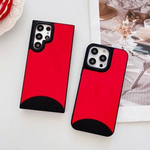 May include: Two red phone cases with black accents. The cases have a rectangular design with a black semi-circle at the bottom. The camera cutouts are visible. The cases are on a white surface.