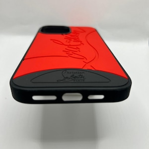 May include: A red and black phone case with the words "Christian Louboutin Paris" embossed on the bottom. The case has a red top with the brand name in a cursive font. The sides and bottom are black.