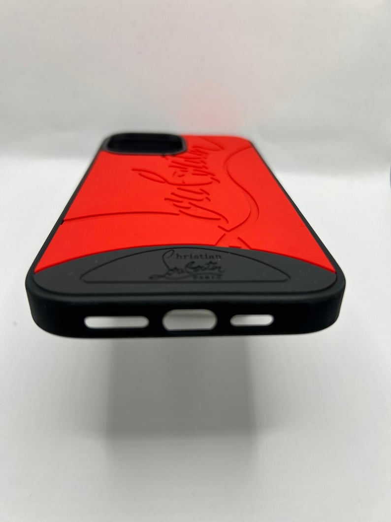 May include: A red and black phone case with the words "Christian Louboutin Paris" embossed on the bottom. The case has a red top with the brand name in a cursive font. The sides and bottom are black.