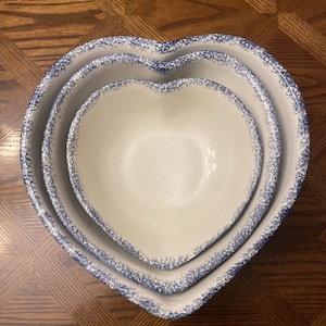 May include: A set of three nested heart-shaped bowls with a cream-colored interior and a blue speckled sponge-painted pattern along the outer rim. The bowls are arranged from largest to smallest, creating a stacked appearance with uniform edges.
