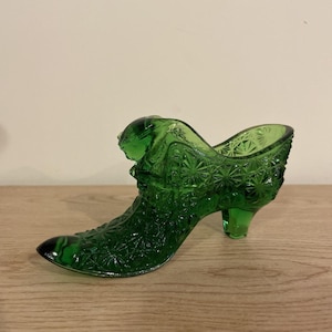 May include: A green glass shoe figurine with a pointed toe and a small heel. The shoe has a detailed, textured surface with a starburst pattern. The decorative shoe is displayed on a wooden surface.