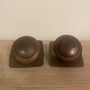 May include: Two cabinet knobs featuring circular wooden rings surrounding dark metallic centers. Each knob is mounted on a square, flat metallic backplate. The wood displays a natural grain texture, while the metal components have a weathered, aged finish.