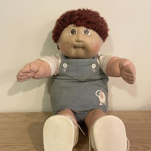 May include: A doll with dark brown yarn hair, a soft vinyl face with rosy cheeks, and brown eyes. The doll wears a white short-sleeved shirt under blue corduroy overalls featuring a small white elephant embroidery, and white fabric shoes with laces.