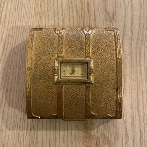 Puede incluir: Un reloj de viaje dorado vintage en una caja rectangular. La esfera del reloj es visible dentro de un marco rectangular más pequeño. La caja tiene una superficie texturizada y detalles metálicos decorativos, que se asemejan a un pequeño baúl.