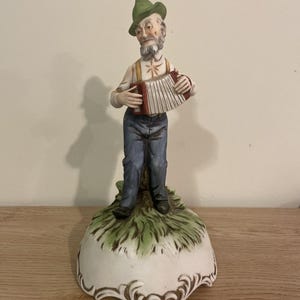 Può includere: Una statuetta in porcellana raffigurante una persona anziana con la barba che indossa un cappello verde, una camicia bianca e pantaloni blu, mentre suona una fisarmonica rossa e bianca. La figura poggia su una base dipinta con erba verde.