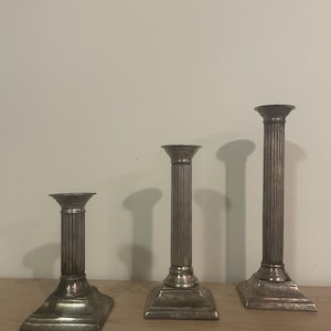 Peut inclure: Trois bougeoirs en métal argenté de hauteurs différentes disposés en rangée. Chaque bougeoir présente une colonne cylindrique cannelée, une base carrée et une coupelle circulaire. La surface métallique offre une finition polie avec des traces d'usure.