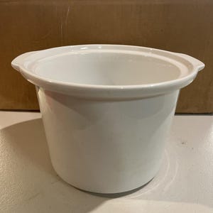 May include: A white ceramic slow cooker insert with a rounded body and a wide rim. The interior is a deep, open bowl shape. The insert is designed to fit inside a slow cooker base for cooking.