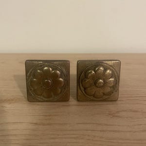 May include: Two square-shaped cabinet knobs made of aged brass metal. Each knob features a raised, circular floral design with eight petals centered on the square face. The metal has a matte, weathered finish with visible dark patina accents.