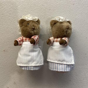 May include: Two brown teddy bear figurines wearing white aprons, red and white checkered shirts, and white bonnets. The bears have small arms and are standing upright. The bottom half of the bears are blue and white striped.