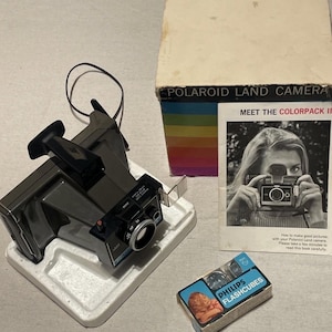 May include: A vintage Polaroid Land Camera, gray in color, with a black viewfinder and lens. The camera sits on a white foam base, next to a box labeled "POLAROID LAND CAMERA" and a booklet. A small box of Philips Flashcubes is also present.
