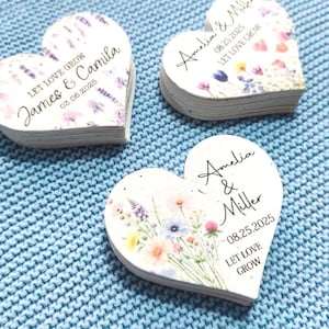 May include: Three heart-shaped wedding favors with floral designs and personalized text. Each favor features names, a date, and the phrase "Let Love Grow." The favors are stacked, with a light blue textured background.