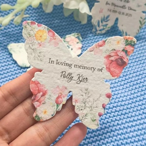 May include: A butterfly-shaped memorial card with pink, yellow, and green floral patterns. The text "In loving memory of Polly Kjer" is printed on the card. Other floral-shaped cards are visible in the background.