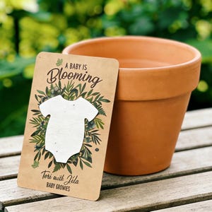 May include: A brown terracotta flower pot beside a seed paper card. The card has a white babygrow shape surrounded by green leaves and the text "A BABY IS BLOOMING." The card also includes the text "Teri with Jela BABY GROWIES."
