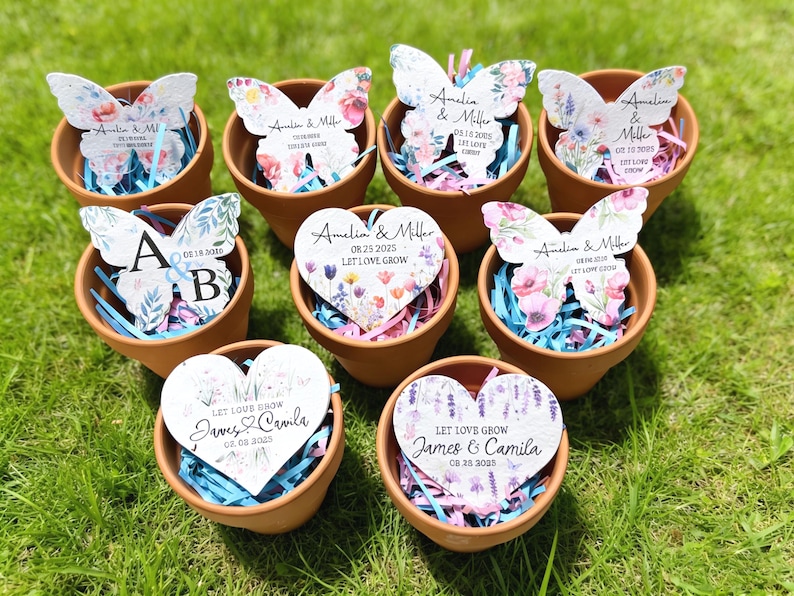 May include: A collection of eight small terracotta pots filled with blue and pink paper shred. Each pot contains a decorative paper cutout in the shape of a butterfly or heart, featuring floral patterns and text including names and dates.