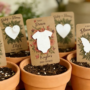 May include: Terracotta pots filled with soil, each holding a baby shower favour. The favours are small cards with a white onesie, heart, or butterfly cut-out, and text that reads "Baby Brown" and "Kayla's Baby Shower."