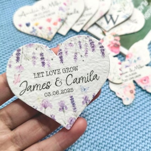 以下が含まれることがあります： 花とラベンダーのアクセントが付いたハート型の結婚式の記念品。「LET LOVE GROW」の文字と「James & Camila」の名前が前面に印刷され、日付は03.06.2025です。他の記念品が背景に見えます。