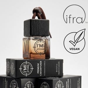 Puede incluir: Una pila de cajas negras y un ambientador para coche. Las cajas dicen "PREMIUM CAR DIFFUSER" y "I'MI STORE". El ambientador tiene una tapa de madera y una botella de vidrio con el logotipo "I'MI STORE". La imagen también incluye el logotipo "VEGAN".