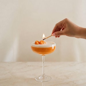 May include: A cocktail glass shaped candle filled with orange wax and topped with a white foam layer and two small orange wax segments. A lit match is held near the candle wick, which is emitting a small flame. The candle is clear glass.