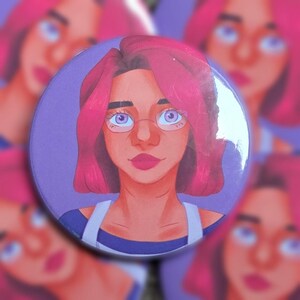 Könnte beinhalten: Ein runder Ansteckbutton mit einer digitalen Illustration einer Person mit leuchtend pinkem Haar, hellvioletten Augen und einer Brille. Die Figur trägt ein dunkelblaues Oberteil mit weißen Trägern vor einem einfarbigen violetten Hintergrund.