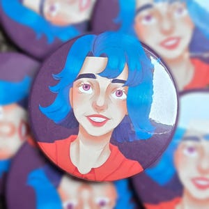 Op de afbeelding: Een ronde button met een gestileerde illustratie van een persoon met felblauw haar en roze ogen. De persoon draagt een rood overhemd met kraag tegen een donkerpaarse achtergrond. Het ontwerp heeft een glanzende afwerking met een zichtbare barst.