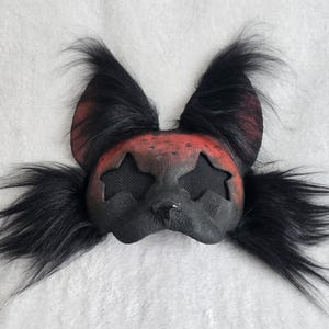 May include: A black and red mask with star-shaped eye cutouts and long, black fur accents. The mask has a textured appearance and is designed to cover the face. The ears are red with black fur.