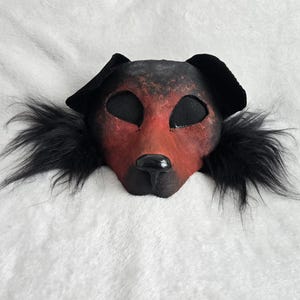May include: A handmade dog mask with a red and black color scheme. The mask features black mesh eye coverings, black ears, and a black nose. The mask is surrounded by black faux fur.