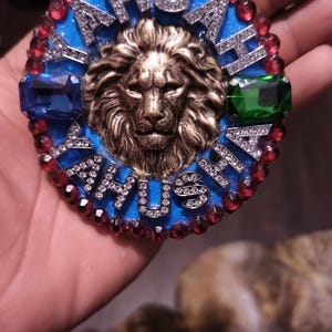 May include: A circular decorative emblem featuring a central bronze-toned lion head relief. The background is bright blue with the text YAHUAH and YAHUSHA spelled out in silver-toned, rhinestone-encrusted letters. Red, blue, and green gems border the piece.