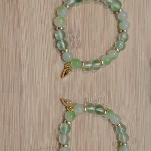 May include: Two beaded bracelets with green and clear beads, accented with gold-colored spacers and a small gold charm. The bracelets are displayed on a light brown wooden surface.