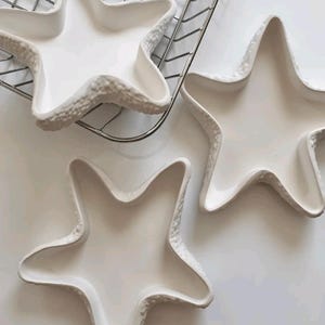 May include: Three white ceramic starfish-shaped dishes. The dishes have a textured, raised edge and a smooth interior. One dish is on a metal wire rack. The dishes are arranged on a white surface.