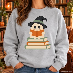May include: A light gray crewneck sweatshirt featuring a graphic of a cream-colored ghost wearing a black witch hat with a green band. The ghost is holding an open orange book while sitting atop a stack of three books in shades of green and orange.