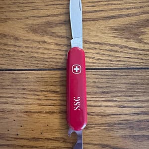 May include: A red multi-tool pocket knife with a single extended silver blade. The handle features a white Swiss cross emblem and the letters SSG printed in white. A metal bottle opener tool is visible at the base of the red handle casing.