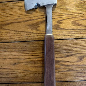 May include: A small hatchet with a polished steel blade and a dark wood handle. The hatchet has a flat top and a curved cutting edge. The handle is rectangular and tapers towards the bottom.