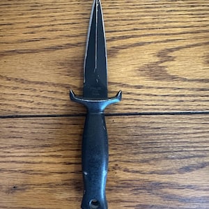 May include: A black dagger with a pointed blade and a handle. The dagger has a guard and a hole at the base of the handle. The blade is black and the handle is a darker shade of black. The dagger is on a wooden surface.