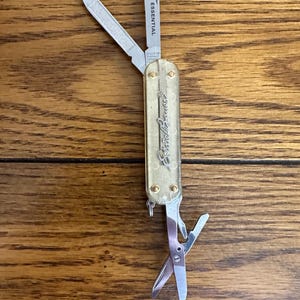May include: A multi-tool pocket knife with a translucent gold handle featuring the Eddie Bauer logo. The tool includes an open blade marked with the word ESSENTIAL, a nail file, and a pair of small scissors extended from the bottom end.