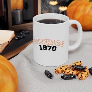 Peut inclure: Tasse en céramique blanche avec l'inscription "VINTAGE 1970" en orange et noir. La tasse contient un liquide foncé et possède une anse incurvée. La tasse est posée sur une surface grise avec des bonbons et des citrouilles d'Halloween en arrière-plan.