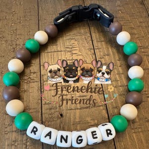 以下が含まれることがあります： 白い文字ビーズで「RANGER」と綴られた犬の首輪。 黒い留め金が付いており、緑、茶色、白のビーズが通されています。 「Frenchie Friends」という言葉も表示されています。