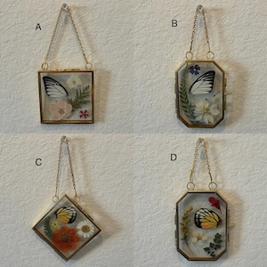 May include: Four decorative glass frames with gold-colored metal edges and hanging chains. Each frame contains a butterfly wing and dried flowers. The frames are different shapes: square, octagonal, and diamond. The butterflies have black, yellow, and white wings.